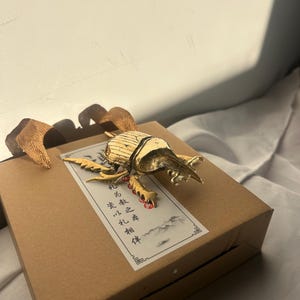 May include: A gold-colored beetle sculpture adorns a brown gift box. The beetle has detailed wings and legs. The box has a decorative label with Chinese characters and a landscape design. The gift box has woven handles.