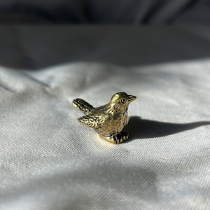 May include: A small, detailed brass bird figurine. The bird is perched, with textured feathers and a slightly open beak. The figurine is a warm gold colour, and sits on a white surface.