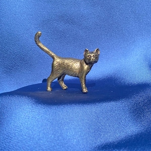 May include: A small, silver-colored figurine of a cat with a curled tail. The cat is standing on a blue surface.