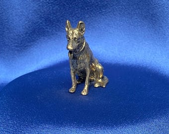 Peut inclure: Une petite figurine en métal dorée représentant un berger allemand assis, la tête légèrement inclinée sur le côté. Le chien porte un collier.
