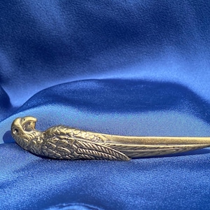 Puede incluir: Un abridor de cartas de metal dorado en forma de pájaro con alas detalladas y una punta afilada.