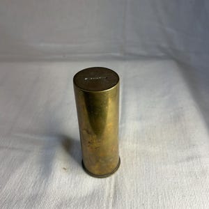 Può includere: Un oggetto cilindrico in ottone con una finitura leggermente ossidata. La parte superiore del cilindro presenta un testo inciso. L'oggetto è in posizione verticale su una superficie bianca. La forma generale è un cilindro alto e sottile.
