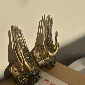 Puede incluir: Dos esculturas de manos doradas con detalles intrincados, montadas sobre bases circulares ornamentadas. Las manos están en un gesto de ofrenda o bendición. Las esculturas están colocadas sobre una superficie marrón, con una etiqueta de papel con caracteres asiáticos.