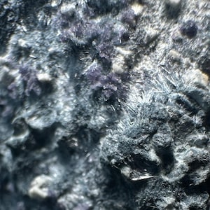 May include: Close-up of a rough, textured rock formation. The surface is primarily dark gray with patches of purple and white crystals. The overall appearance is rugged and natural, with a variety of textures.