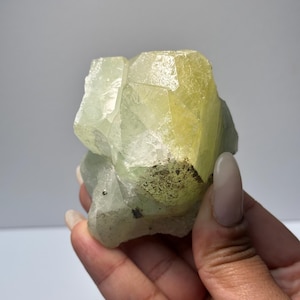 May include: A light green and yellow crystal cluster with a faceted, geometric structure. The crystal has a translucent quality, with variations in color and texture. The crystal is held in a hand.