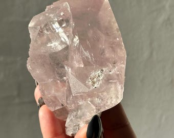 May include: A light pink, crystalline mineral specimen with a rough, irregular shape. The crystal is held in a hand with black nail polish. The mineral has a translucent quality, with some areas appearing more opaque.