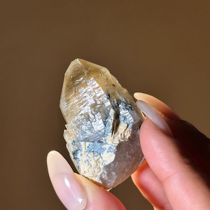 May include: A clear quartz crystal with a pointed tip and a rough, textured base. The crystal has a light brown hue with areas of blue and white. The crystal is held in a hand with a neutral background.
