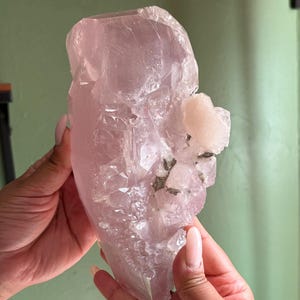 May include: A large, translucent, light pink crystal cluster with a rough, irregular shape. The crystal has a smooth, polished surface in some areas and a more textured, crystalline structure in others. A smaller, off-white crystal formation is attached to the main crystal.