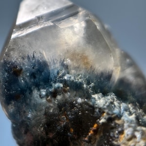 May include: Close-up of a clear quartz crystal with blue inclusions. The crystal has a pointed tip and a rough, textured base with hints of orange and brown. The blue inclusions appear as small, needle-like formations within the clear quartz.