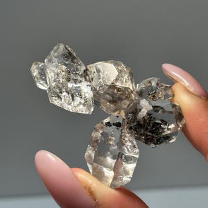 Peut inclure: Un amas de cristaux de quartz transparents à facettes multiples présentant des inclusions internes sombres d'aspect métallique. Le spécimen minéral présente des bords géométriques nets et une surface transparente très brillante.