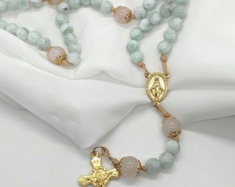 May include: A rosary with pale green and pink beads. The beads are strung on a light brown cord, with gold-colored metal accents, including a crucifix and a pendant with a religious figure. The background is a white fabric.