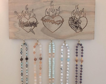 Può includere: Organizzatore da parete in legno per gioielli con tre immagini incise di cuori fiammeggianti. I cuori sono adornati da spine, una spada e gigli. Sei collane di perline pendono da ganci sotto le immagini.