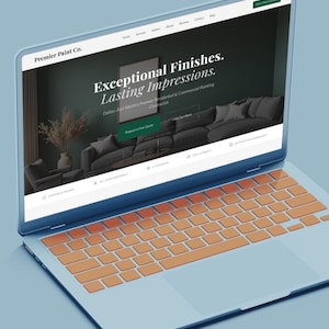 May include: A light blue laptop displaying the Premier Paint Co. website. The website features the text "Exceptional Finishes. Lasting Impressions." The keyboard has orange keys. The screen shows a dark green background with a sofa and a framed picture.