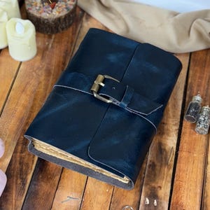 Puede incluir: Un diario de cuero azul oscuro con cierre de hebilla de latón. El diario tiene páginas envejecidas y amarillentas y una correa de cuero. El fondo incluye velas decorativas, cristales y pequeños frascos de vidrio sobre una superficie de madera.