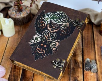 May include: A brown leather journal with an owl and Celtic knot design. The journal has metal clasps and is surrounded by decorative items, including crystal trees, candles, and gemstones. The journal is on a wooden surface.