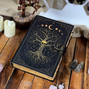 May include: A black journal with a gold tree of life design and a skull. The cover features a crescent moon and stars. The journal is closed with a metal clasp. The page edges are gold.