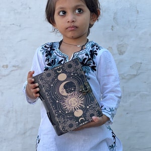 May include: A young person wearing a white embroidered top holds a black leather journal with a sun and moon design. The journal has a clasp and a decorative metal accent.