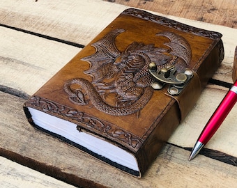 May include: A brown leather journal with a dragon design embossed on the cover. The journal has a metal clasp and is closed. A red pen is lying on a wooden surface next to the journal.