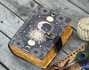 Peut inclure: Un journal relié en cuir noir avec un motif céleste représentant un soleil, une lune et des étoiles. Le journal est doté d'un fermoir en laiton et d'une sangle en cuir noir.