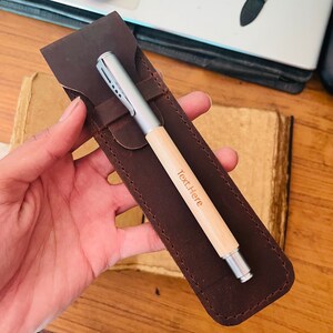 Op de afbeelding: Een bruin leren pennenetui met een zilveren en houten pen erin. De pen heeft de tekst "Text.Here" gegraveerd op het hout.