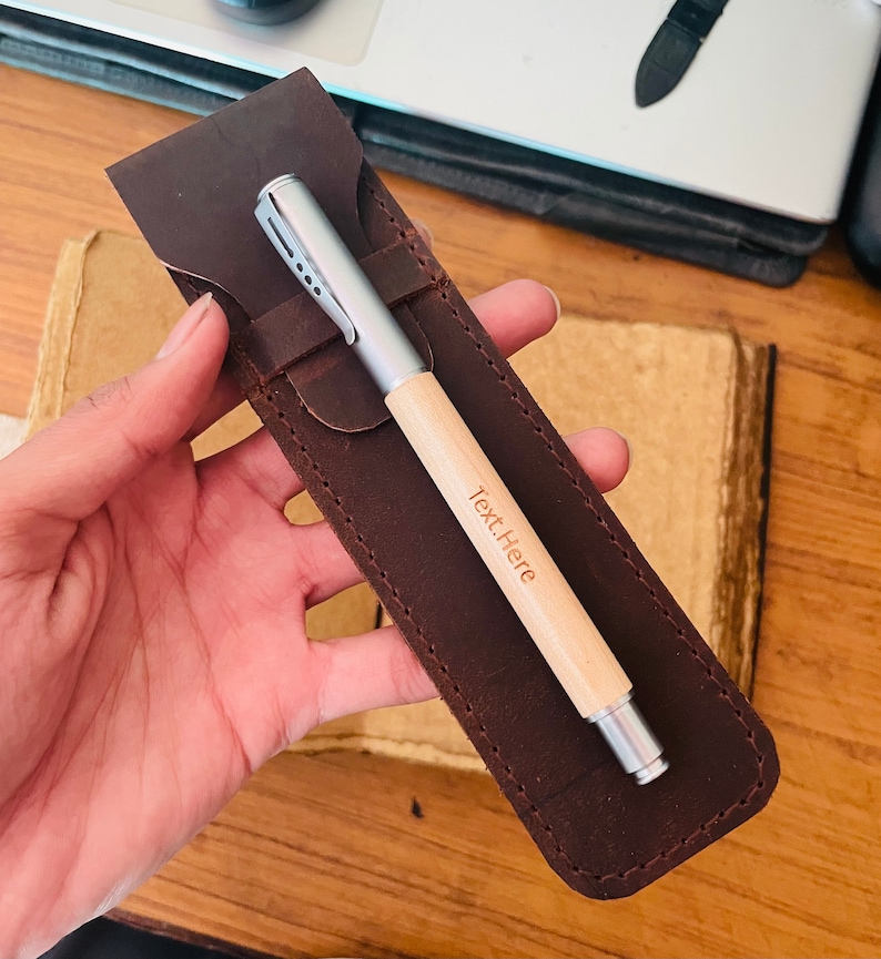 Op de afbeelding: Een bruin leren pennenetui met een zilveren en houten pen erin. De pen heeft de tekst "Text.Here" gegraveerd op het hout.
