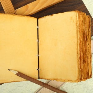 May include: A blank, yellowed, leather-bound journal with a black ribbon closure. The journal is open and lying flat on a light green surface. A pencil is lying on top of the journal.