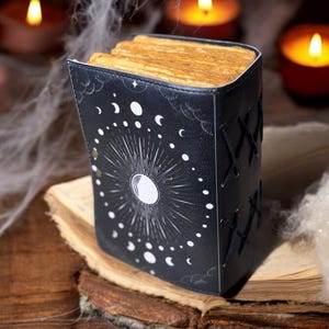 May include: A black leather-bound journal with a moon and stars design. The journal is closed and sitting on an old book with a worn, yellowed page.