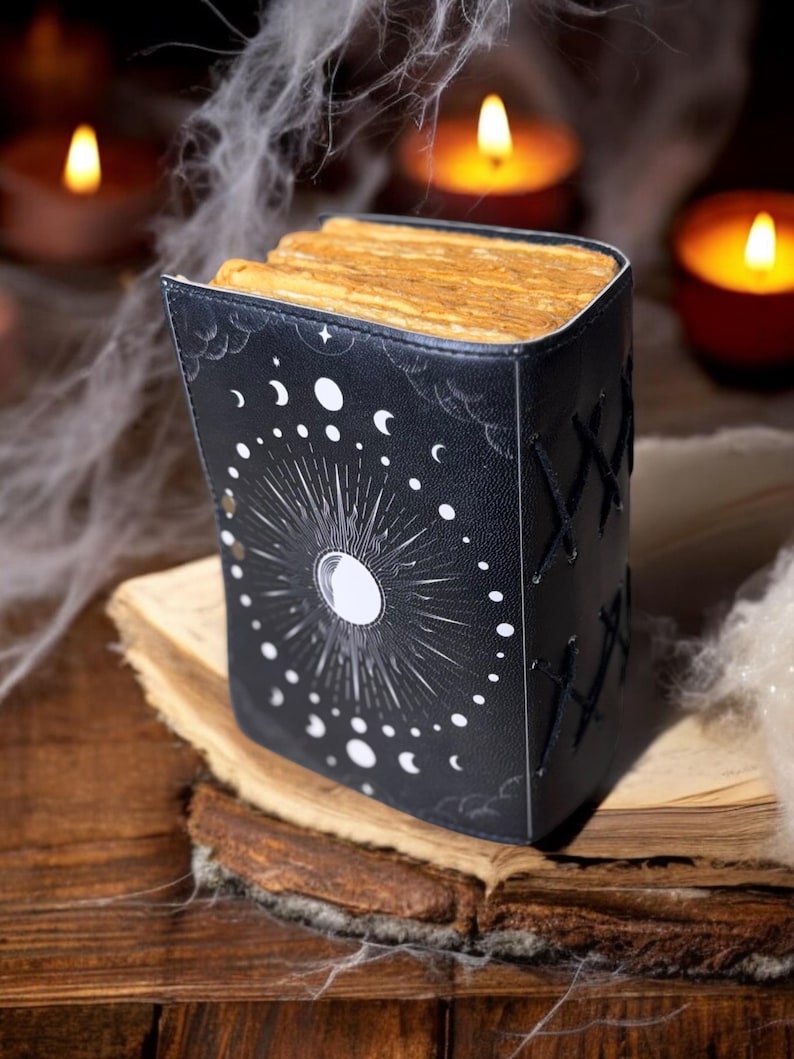 May include: A black leather-bound journal with a moon and stars design. The journal is closed and sitting on an old book with a worn, yellowed page.