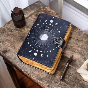 May include: A black leather-bound journal with a celestial design featuring a sun, moon, and stars. The journal has a metal clasp and is closed.