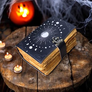 May include: A black leather-bound journal with a moon and stars design. The journal has a clasp closure and aged pages. The journal is sitting on a wooden surface with candles.