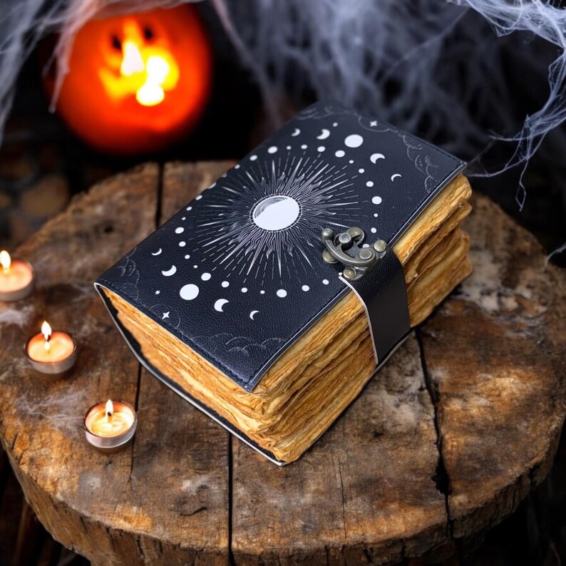 May include: A black leather-bound journal with a moon and stars design. The journal has a clasp closure and aged pages. The journal is sitting on a wooden surface with candles.