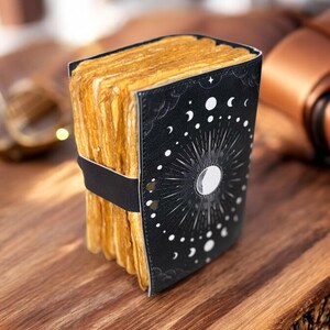 May include: A black leather-bound journal with a moon and stars design. The journal is closed and has a band around it. The journal is sitting on a wooden surface.