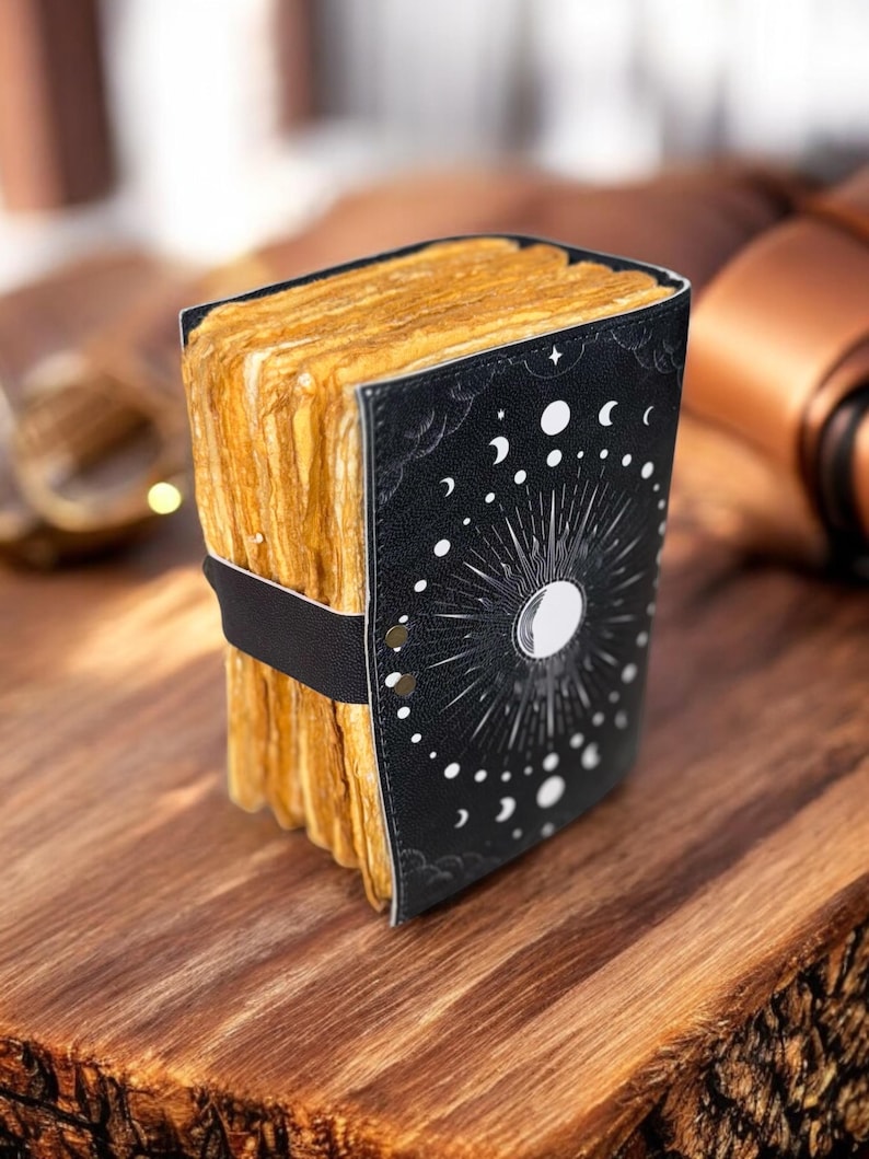 May include: A black leather-bound journal with a moon and stars design. The journal is closed and has a band around it. The journal is sitting on a wooden surface.