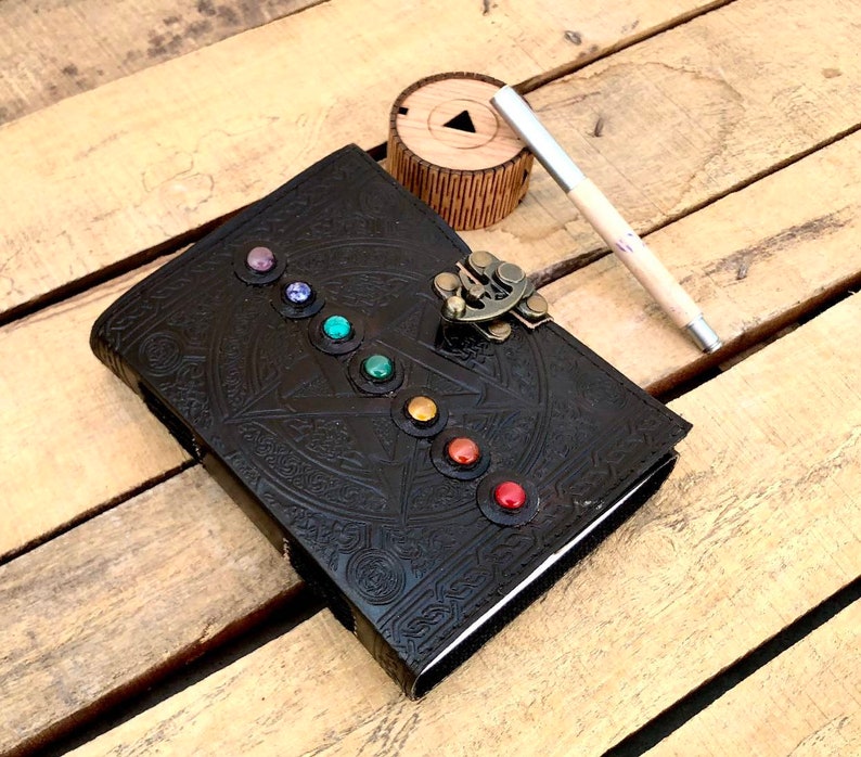 May include: A black leather journal with a decorative embossed design and a metal clasp. The journal has a row of colorful gemstones around the edge.