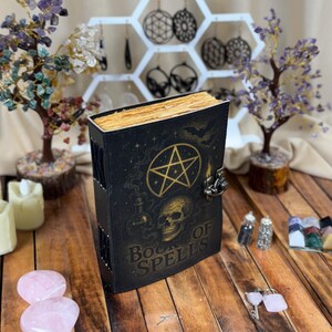 May include: Black "Book of Spells" with gold accents, a pentagram, and a skull. The book has a metal clasp and aged-looking pages. The cover also features bats and potion bottles. The book is on a wooden surface.
