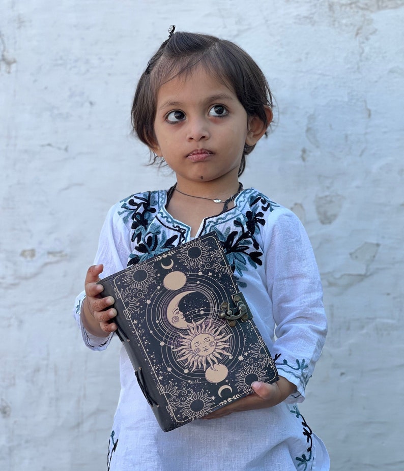 May include: A young person wearing a white embroidered top holds a black leather journal with a sun and moon design. The journal has a clasp and a decorative metal accent.
