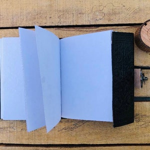May include: A black leather-bound journal with blank pages, open to reveal the white paper inside. A wooden pen holder and a pen are on a wooden surface.