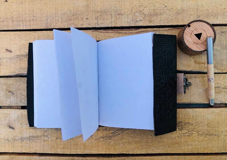 May include: A black leather-bound journal with blank pages, open to reveal the white paper inside. A wooden pen holder and a pen are on a wooden surface.