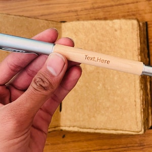 Op de afbeelding: Een zilveren en houten pen met de tekst "Text.Here" gegraveerd op het hout. De pen wordt in een hand gehouden.