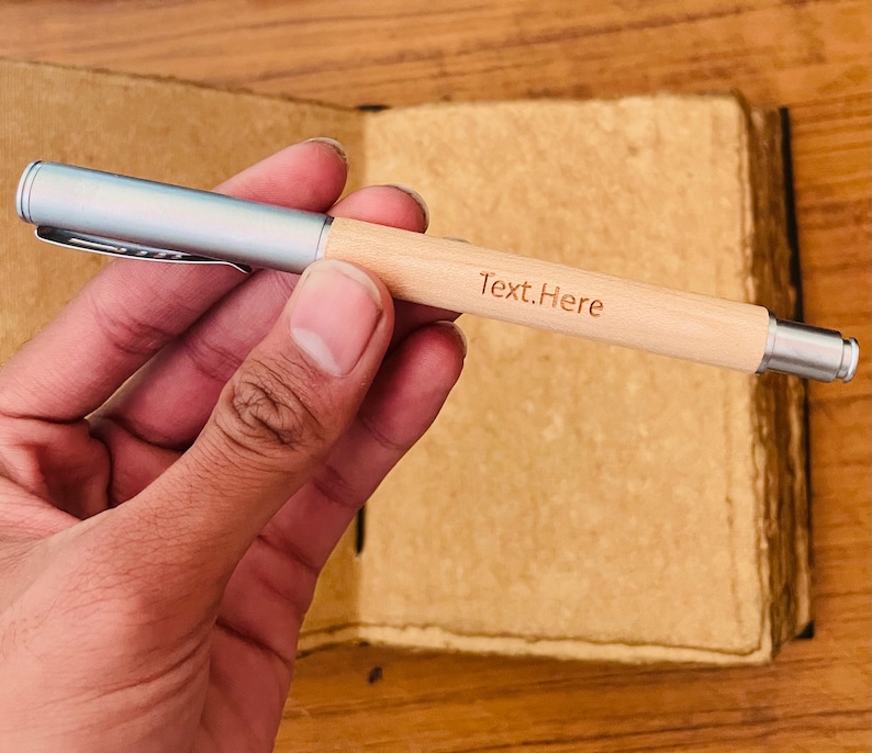 Op de afbeelding: Een zilveren en houten pen met de tekst "Text.Here" gegraveerd op het hout. De pen wordt in een hand gehouden.