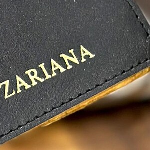 May include: Close-up of a black leather wallet with gold stitching and the name "Zariana" embossed in gold lettering.