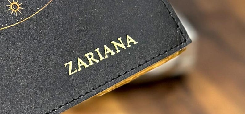 May include: Close-up of a black leather wallet with gold stitching and the name "Zariana" embossed in gold lettering.