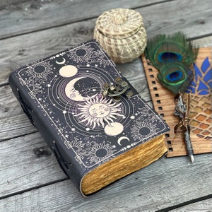 May include: A black leather journal with a celestial design featuring a sun, moon, and stars. The journal has a clasp closure and is decorated with a decorative metal piece. The journal is open to reveal blank pages.