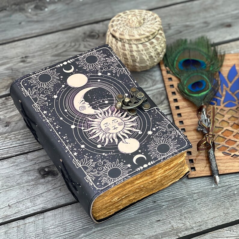 May include: A black leather journal with a celestial design featuring a sun, moon, and stars. The journal has a clasp closure and is decorated with a decorative metal piece. The journal is open to reveal blank pages.