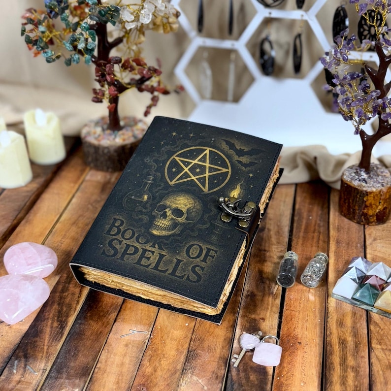 May include: A black leather-bound book titled "Book of Spells" with gold embossed designs, including a skull, pentagram, and bottles. The book has a metal clasp and aged-looking pages. Decorative items like crystals and miniature trees surround the book.