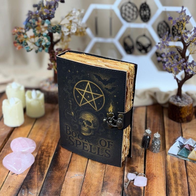 May include: A black leather-bound book with the words "Book of Spells" in gold lettering. The cover features a gold pentagram, skull, and other occult symbols. The book is secured with a black strap and metal clasp. The pages have a vintage, aged appearance.