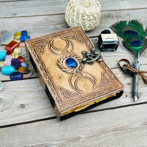 Op de afbeelding: Een bruin leren dagboek met een drievoudig maandesign en een blauwe edelsteen. Het dagboek heeft een metalen sluiting en is versierd met Keltische knopen.