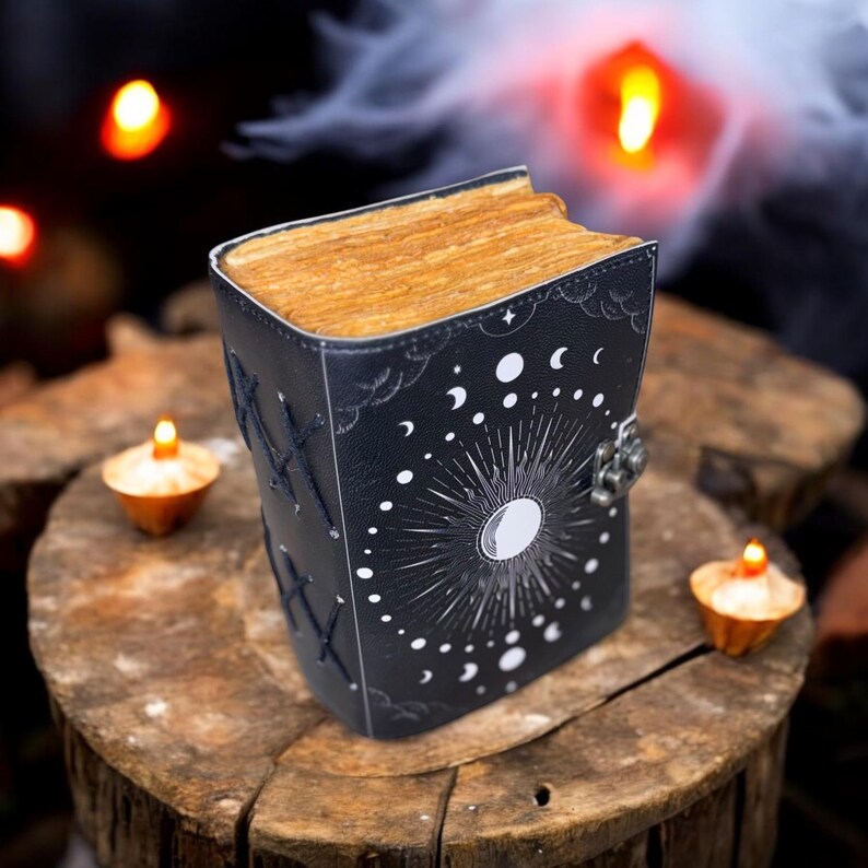 May include: A black leather-bound journal with a moon and stars design. The journal has a silver clasp and is closed. The journal is sitting on a wooden surface with candles in the background.