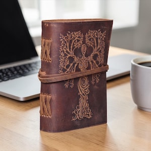 May include: A dark brown leather journal with a tree of life design embossed on the cover. The journal is closed with a leather strap and has stitched detailing on the spine. A laptop and a mug of coffee are in the background.