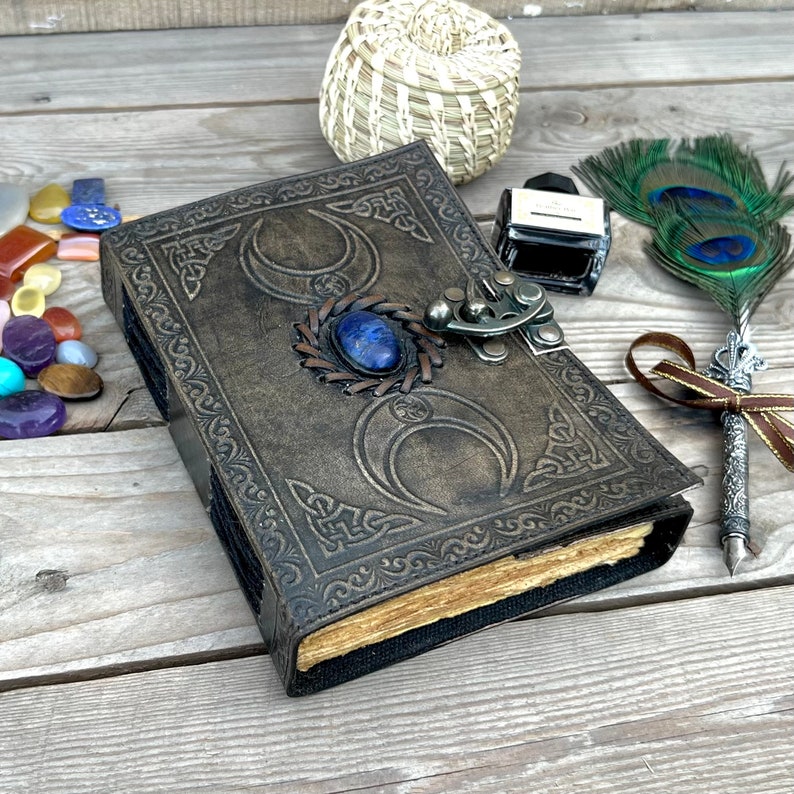 Op de afbeelding: Een bruin leren dagboek met een decoratief reli&euml;fpatroon met drie halve manen en een blauwe edelsteen. Het dagboek heeft een metalen sluiting en is gesloten.